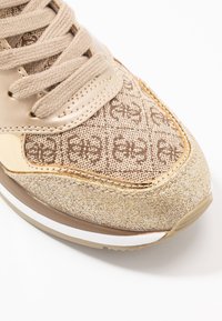 Beige lenkkarit, joissa on teksturoitu kankainen päällinen ja kudottu kuvio, metalliset yksityiskohdat ja kiiltävä pinta; ulkopohjassa on valkoista ja kultaa.