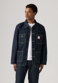 Mørk denimjakke med knaplukning, to store forlommer og kontrastfarget hvit søm. Båret over en hvit t-skjorte.