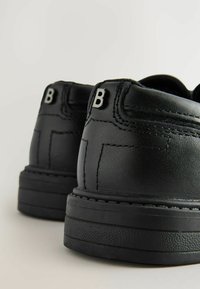 Scarpe in pelle nera con design alto, dotate di cuciture a contrasto, una suola testurizzata e un accent principale in metallo a forma di lettera "B" sul retro.