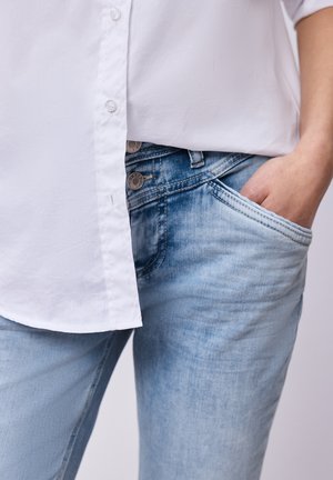 Osoba nosząca wyblakłe niebieskie dżinsy z ręką w kieszeni oraz białą koszulę na guziki, wpuszczoną z przodu.