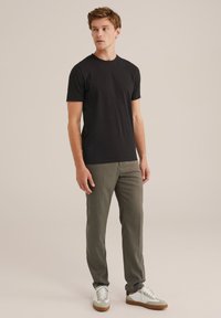 Joven vestido con una camiseta negra de manga corta, pantalones grises y zapatillas blancas, de pie contra un fondo beige liso.