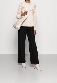 Cremetöniger, rüschierter Button-Up-Hemd, schwarze, weit geschnittene Hose, beigefarbene, hohe Stiefeletten und eine gemusterte Stofftasche mit neutralem Design.
