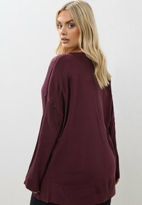 Blond kvinna som ser över axeln iklädd en lös vinröd tröja mot en enkel vit bakgrund.