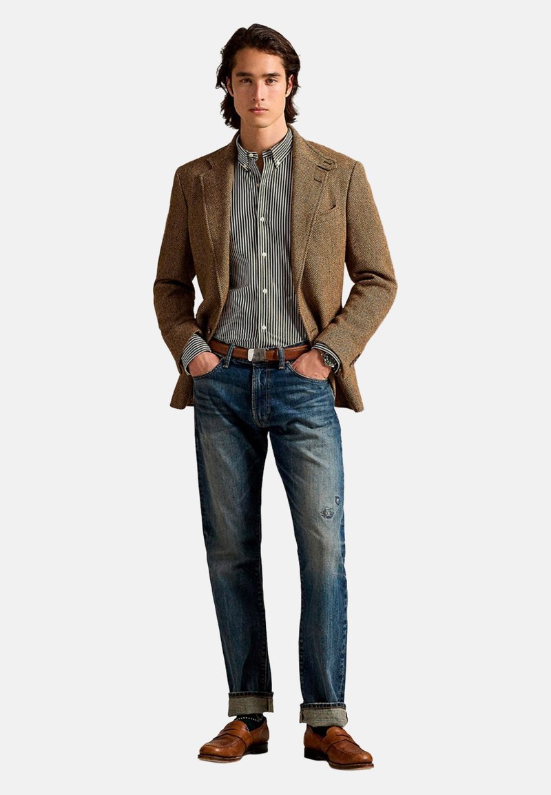 Veste en tweed marron portée sur une chemise rayée, associée à un jean bleu délavé et des chaussures en cuir marron. Détails comprenant une ceinture et des manches retroussées.