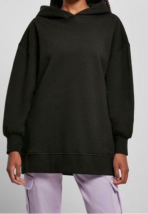 Osoba nosząca za dużą czarną bluzę z kapturem z długimi rękawami i prążkowanymi mankietami, połączoną z jasnofioletowymi spodniami typu cargo na szarym tle.