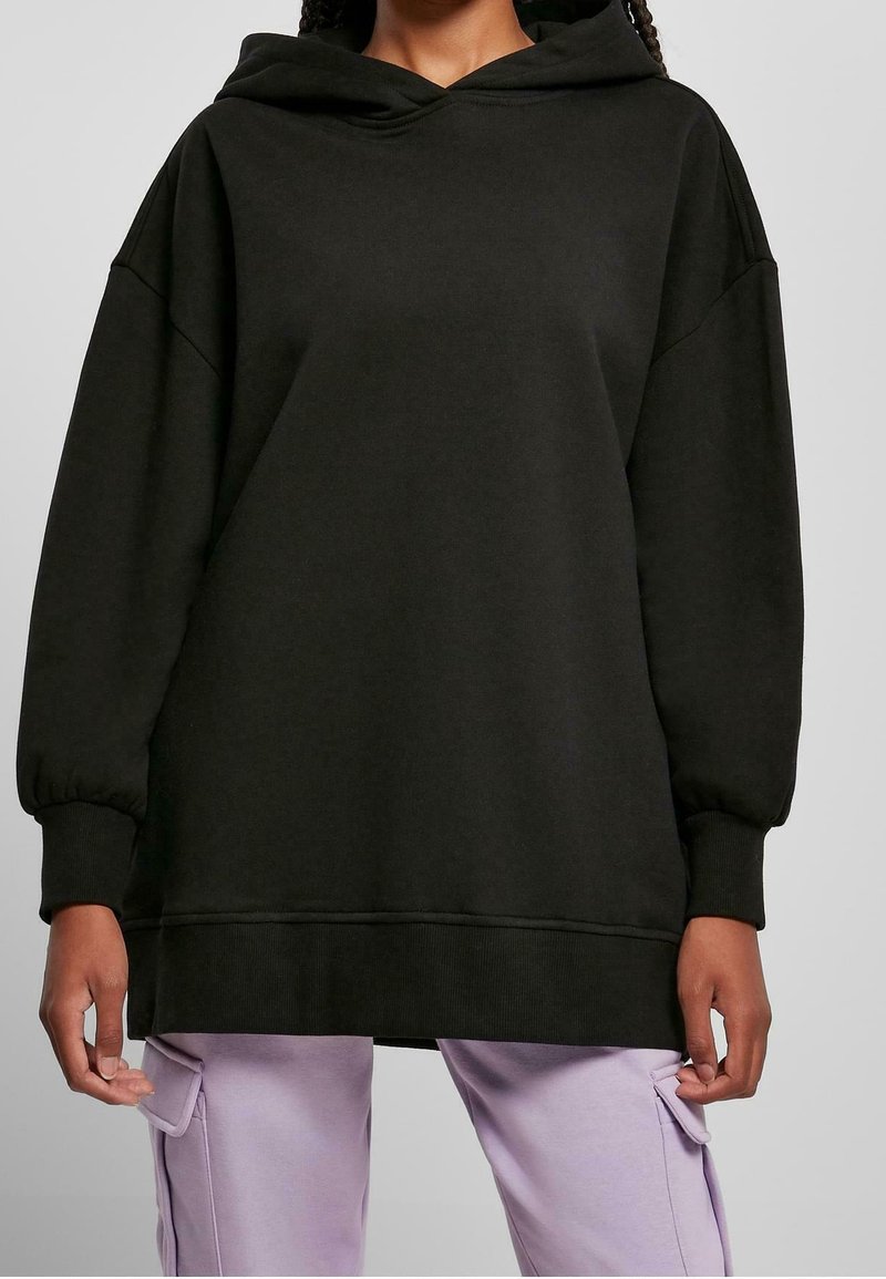 Personne portant un sweat à capuche noir oversize avec des manches longues et des poignets côtelés, associé à un pantalon cargo violet clair sur un fond gris.