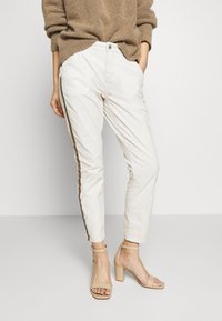 Ljusbeiga, avsmalnande byxor med brun sida, med framfickor och knappstängning, tillsammans med beiga högklackade sandaler.