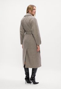 Abrigo de mezcla de lana beige con mangas largas, cintura ceñida con cinturón y una abertura en la parte trasera para facilitar el movimiento, combinado con botas negras de tacón alto y punta. 