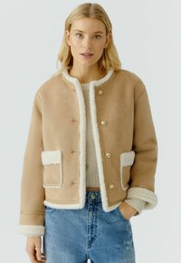 Beige jas met witte nepbont afwerking, ronde kraag, knopen aan de voorkant en twee opgestikte zakken. Getextureerde stof met een gladde afwerking.
