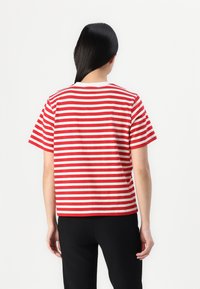 Άτομο με μακριά μαύρα μαλλιά που φοράει κοντομάνικο κόκκινο και λευκό ριγέ πουκάμισο και μαύρο παντελόνι, εμφανιζόμενο από πίσω σε λευκό φόντο.