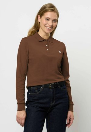 Femme souriante portant un polo marron à manches longues avec des boutons et un jean bleu foncé avec une ceinture noire sur un fond uni.