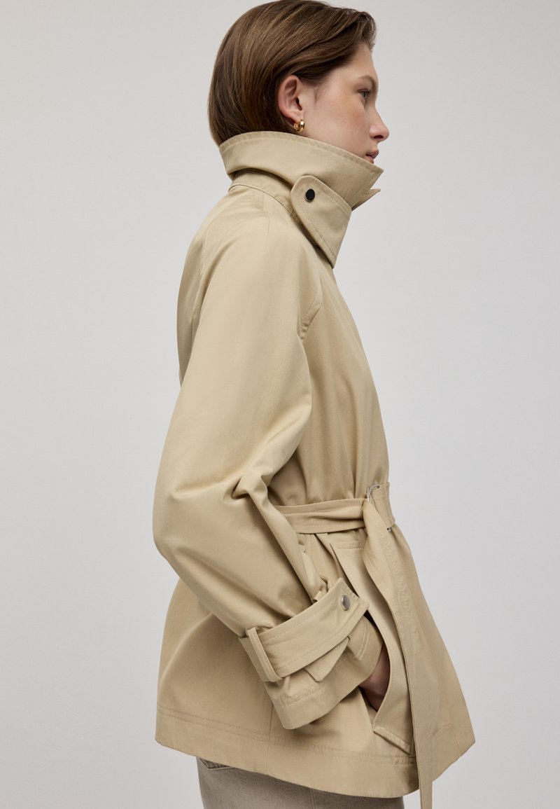Beige wasserdichte Jacke mit hohem Kragen, tailliertem Schnitt und Seitentaschen. Verfügt über Knopfmanschetten und eine glatte Textur.