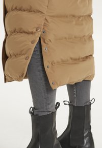 Brun pufferjakke med snaplukning i siden, parret med grå jeans og sorte ankelstøvler med trækflapper og strukturerede paneler.