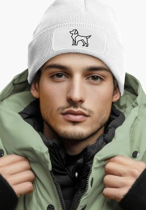 White knitted beanie with a rectangular patch featuring a black line drawing of a dog. Worn with a green padded jacket over a black layer.
