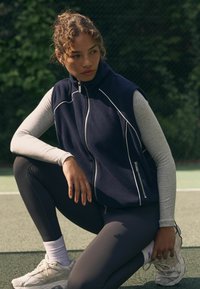 Tumesinine fliisvest kõrge krae, tõmbluku ja valgete triipudega. Hall pika varrukaga särk, mustad retuusid ja heledad spordijalatsid. Välitingimustes.