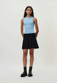 Kvinne med langt krøllete hår som har på seg en ermeløs lyseblå topp, svart svingkjole og svarte ankelboots, står med hendene bak ryggen.