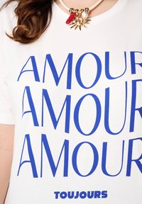 Witte T-shirt met vet blauwe tekst die "AMOUR" meerdere keren spelt. De halslijn heeft een chunky decoratieve ketting met bedels.