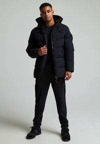 Sort pufferjakke med lynlås foran, hætte og polstret design. Kombineret med sorte bukser og mørke sneakers. Minimalistiske detaljer, glat tekstur.