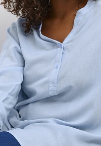 Frau in einer hellblauen, knöpfbaren Bluse mit lockigem Haar, gezeigt von Hals bis Taille, vor einem einfarbigen hellen Hintergrund.
