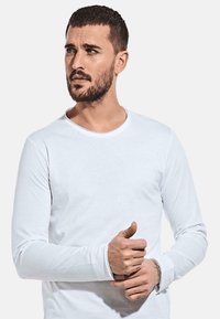Weißes Langarmshirt aus glattem Baumwollstoff mit rundem Ausschnitt und eng anliegenden Bündchen. Das Modell steht mit neutralem Gesichtsausdruck.