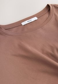 Brun bomulls-T-shirt med slät yta, rund halsringning och synliga sömmar; har en varumärkesetikett med texten "NEXT" på insidan.