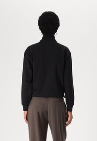 Pull noir à col montant, avec un motif texturé et des manches longues. Associé à un pantalon gris, avec un détail de poche arrière.