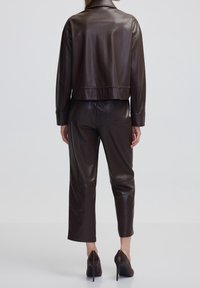 Giubbotto corto in pelle marrone con colletto alto e cuciture decorative abbinato a pantaloni in pelle marrone. Presenti scarpe con tacco alto.