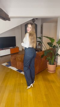 Temno modre kavbojke širokih nogavic, kombinirane z belo črtasto majico z dolgimi rokavi, postavljene v moderno dnevno sobo z notranjimi rastlinami.