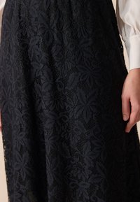 Jupe en dentelle noire avec des motifs floraux portée par-dessus une doublure claire, accompagnée d'une chemise à manches longues blanche et d'une main posée sur le côté.