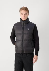 Gilet imbottito nero con collo alto, chiusura zip, design trapuntato e accento del logo sul petto, indossato sopra una maglietta nera a maniche lunghe.