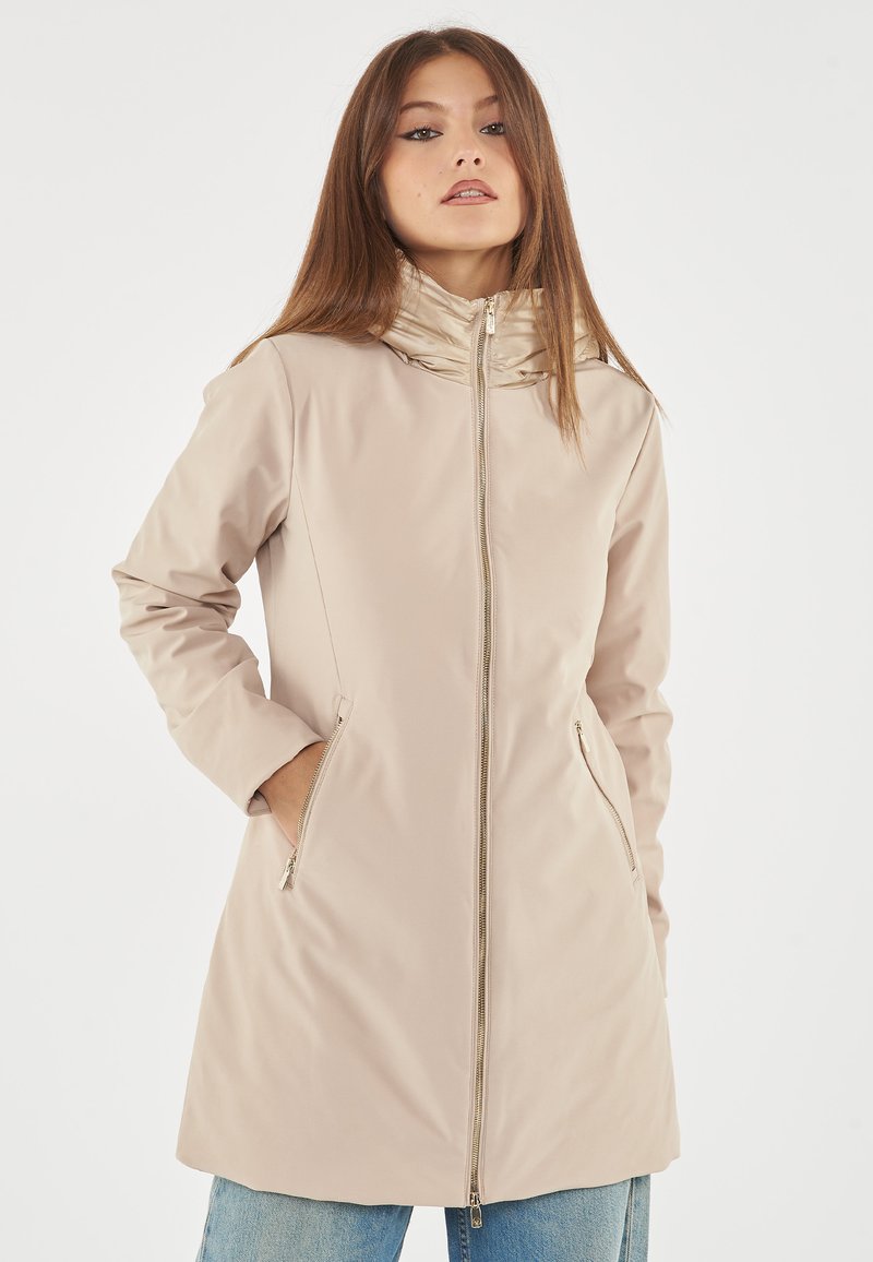 Cappotto impermeabile beige con colletto alto, zip frontale e tasche laterali. Texture liscia e silhouette aderente con maniche lunghe.