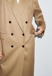 Double-breasted coat in beige fabric with a textured finish. Features black buttons and side pockets, with wide lapels and tailored design.