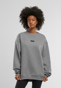 Felpa grigia oversize con spalle basse, polsini e orlo a coste. Presenta una piccola patch rettangolare nera con testo bianco.