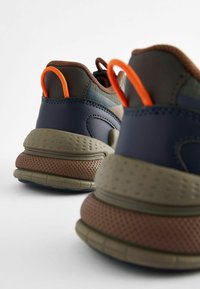 Die Sneakers bestehen aus einer Mischung von Materialien in Dunkelgrün, Marineblau und Braun. Sie sind mit einer orangen Zuglasche und strukturierten Sohlen für besseren Halt akzentuiert.