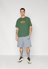 Grøn bomulds t-shirt med et gult logo, parret med grå sporty shorts. Sorte sneakers og grå sokker med Nike-logoen.