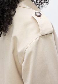 Cheveux bouclés et foncés reposant sur une épaule en tissu beige avec une épaulette à bouton sur un vêtement, mettant en valeur les détails de couture et de texture.