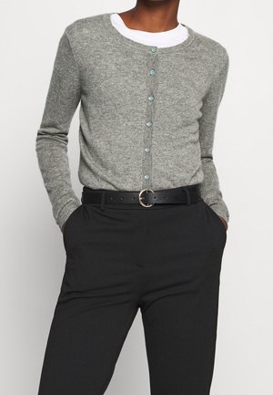 Person trägt eine graue geknöpfte Strickjacke über einem weißen Hemd, das in eine schwarze Hose gesteckt ist, ergänzt durch einen schwarzen Gürtel mit einer silbernen runden Schnalle.