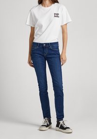 Camiseta blanca de manga corta con logo en el pecho, combinada con jeans skinny de color azul oscuro y zapatillas altas negras con suelas blancas.