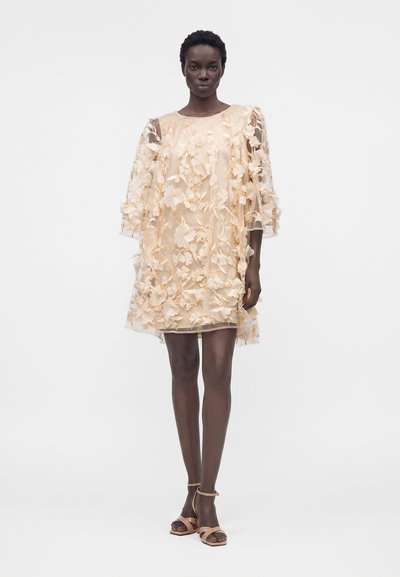 Γυναίκα που φορά μακριά μέχρι το γόνατο διάφανη μπεζ φορεσιά διακοσμημένη με 3D λουλουδένιες εφαρμογές και μπεζ σανδάλια με τακούνι, στέκεται σε λευκό φόντο.