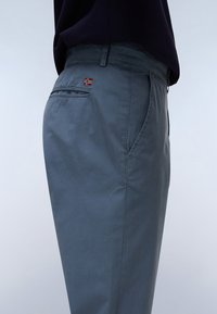 Grå bukser med en glat tekstur, der har en baglomme med en lille broderet flagdetalje. Lige ben-design, afslappet stil.