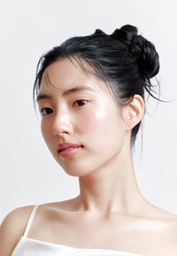 Carnagione chiara con una leggera macchia rosa sulla guancia inferiore sinistra. Capelli neri raccolti in uno chignon, indossa una canottiera bianca con spalline sottili su uno sfondo semplice.