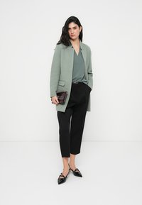 Ανοιχτό πράσινο σακάκι πάνω από γκρι τοπ, μαύρο παντελόνι και μαύρα μυτερά παπούτσια. Κρατά μικρό μπορντό clutch. Καθαρή, μοντέρνα σιλουέτα.