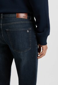 Vaqueros de mezclilla oscura con un parche de cuero marrón en la cinturilla que dice "BOSS". Presenta un bolsillo trasero con un acento de logo metálico. Vista de cerca.