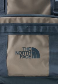 Μπεζ και μπλε υφασμάτινη θήκη σακιδίου με οριζόντια λουριά και το λογότυπο "The North Face" τυπωμένο σε σκούρο μπλε.