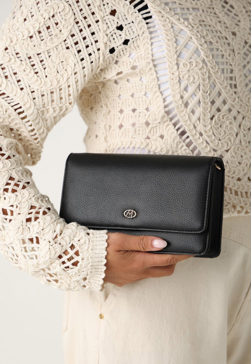 Portafoglio in pelle nera con texture a pallini, dotato di logo dorato e chiusura a patta, tenuto in mano vicino a una manica bianca all'uncinetto.