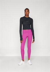 Fekete, hosszú ujjú felső ívelt hemvonalú és logóval, testhezálló rózsaszín leggings-szel és fehér sportcipőkkel párosítva. Sima szövettextúra.