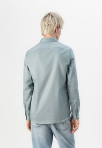 Camisa de manga larga de color azul claro con cuello, puños de botón y textura suave. Combinada con jeans de mezclilla claros. Vista trasera, mostrando la longitud de la camisa.