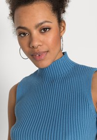 Débardeur à col roulé sans manches en bleu doux, présentant un col haut et une texture subtile. Accentuée par une silhouette ajustée.