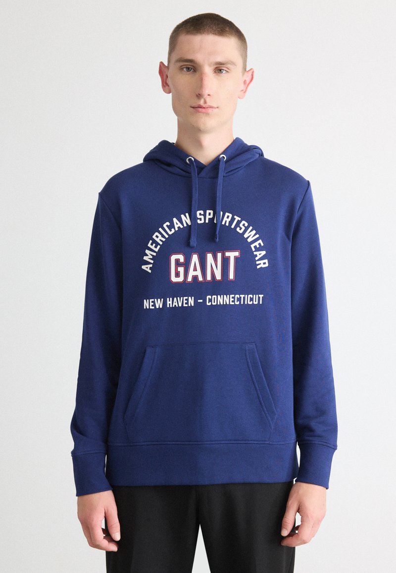 Sudadera navy con cordón, que presenta un bolsillo tipo canguro en la parte frontal y texto impreso: "AMERICAN SPORTSWEAR GANT NEW HAVEN - CONNECTICUT."