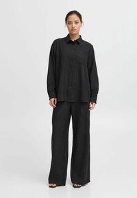 Zwarte button-up shirt met een enkel zakje, gecombineerd met een broek met een brede pijp. De stof heeft een subtiel textuurpatroon.